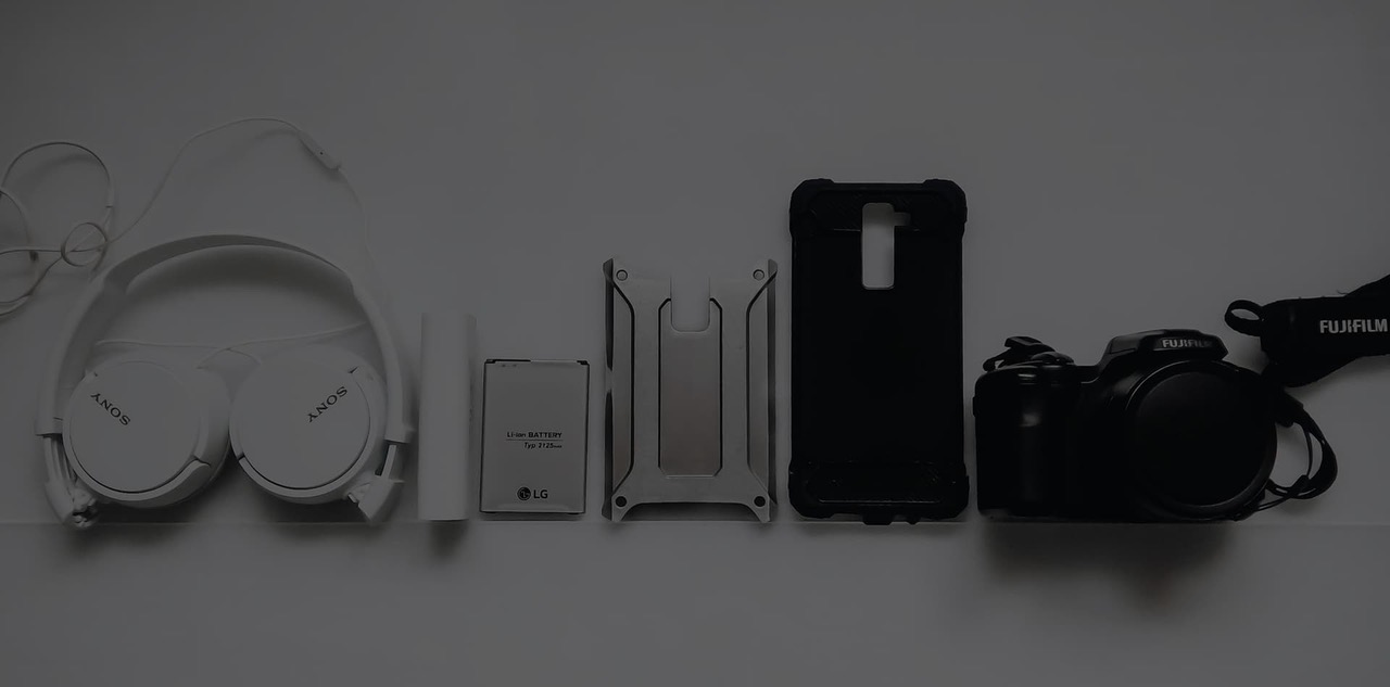 Over-ear headphones, a spare battery, two phone cases and a compact camera lined up evenly on a pale studio surface