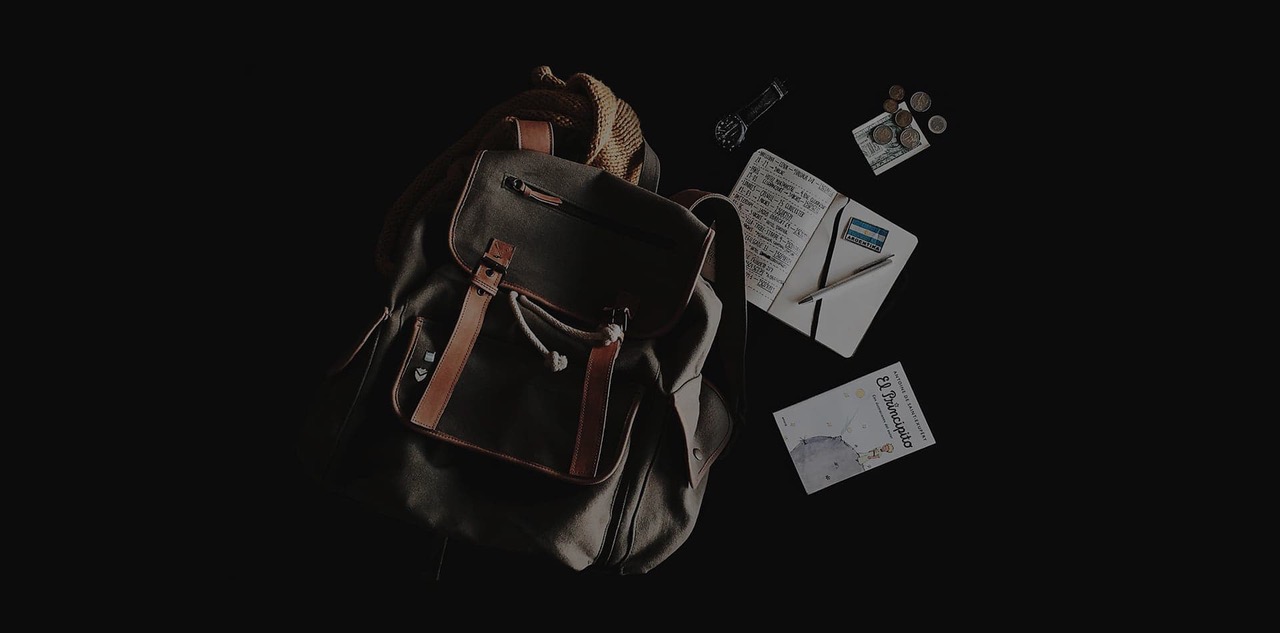 Canvas and leather rucksack on a dark surface beside a handwritten notebook, pen, watch, coins, folded cash and a paperback
