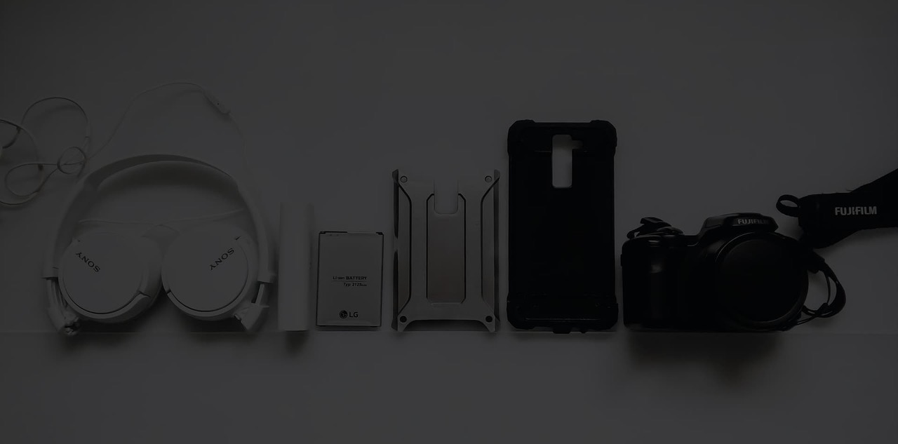 The same tech accessory lineup shown wider and darker: headphones, battery, phone cases and camera evenly spaced on a grey surface